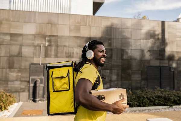 African american deliveryman in special yellow uniform, carrying orders in backpack for customers and holding cardboard box while listening music via bluetooth earphones