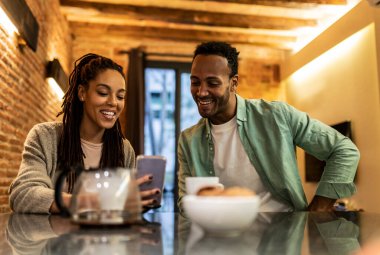 Neşeli Afrikalı kız arkadaş yemek odasında erkek arkadaşına cep telefonunu gösteriyor. Onlar evde birlikte boş zaman geçiriyorlar. - Kadınlara odaklan. -