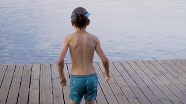 Mały chłopiec w kombinezonie skaczący do jeziora z drewnianego molo. Zabawa w letni dzień. — Wideo stockowe