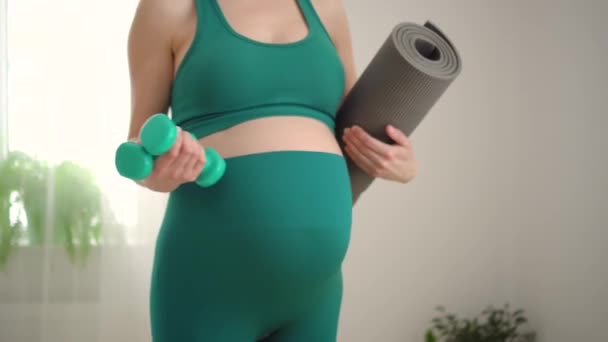 Egy terhes nő súlyzókkal edz. Fiatal lány kezében egy jóga matrica. — Stock videók