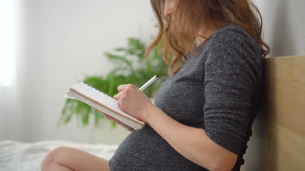 Schwangere macht sich Notizen in Notizbuch, macht Liste der notwendigen Dinge für Schwangerschaft, Geburt — Stockvideo