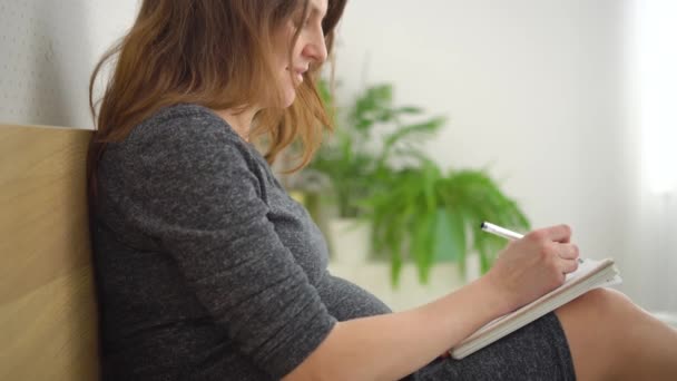 Беременная женщина делает заметки в блокноте, составляет список необходимых вещей для беременности, родов — стоковое видео