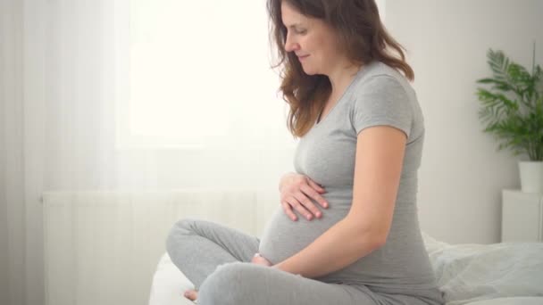 귀여운 임산부가 침대에 앉아 있습니다. 행복 한 임신이라는 개념. 9 개월 간 아기를 기다리며. — 비디오