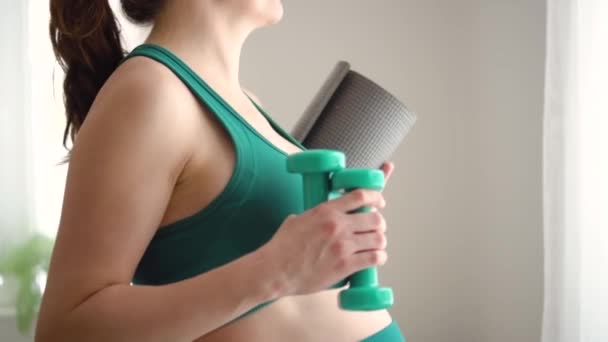 Een zwangere vrouw bereidt zich voor op het trainen met halters. jong meisje met een yoga mat in de hand. — Stockvideo