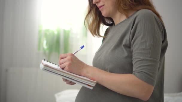 Gravid kvinna antecknar i en anteckningsbok, gör en lista över nödvändiga saker för graviditet, förlossning — Stockvideo