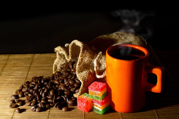 Kaffeebohnen aus dem Jutesack geschüttet, orangefarbene Tasse Kaffee und bunte Gelee-Bonbons auf Holzmatte — Stockfoto