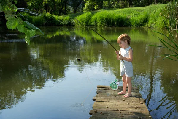 Cute Little Boy Pesca a partir da borda da doca de madeira e esperando pacientemente por sua captura no dia ensolarado — Fotografia de Stock
