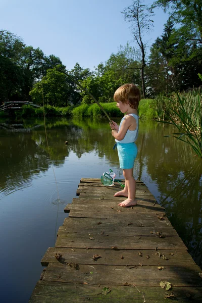 Süßer kleiner Junge steht am Rand des hölzernen Docks und angelt am See bei sonnigem Tag — Stockfoto