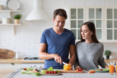 Beautiful young couple smiling while preparing vegetable salad in the kitchen at home. Healthy and wholesome food.