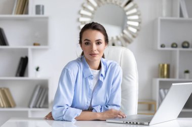 Business woman in a blue shirt at home office with a laptop look at camera.