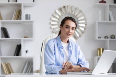 Business woman portrait in blue shirt at home office with laptop looking at the camera.