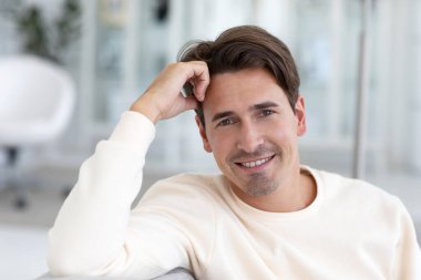 Yakışıklı, sakin bir adamın yakın plan portresi çekici bir görünüme sahip, rahat bir iç mekâna karşı kanepede oturuyor. Öğrenci içeride poz veriyor.