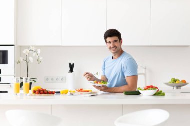 Yakışıklı genç adam öğle yemeği arasında sağlıklı yiyecekler yiyor tabakla birlikte mutfağın içinde açık renkli sebzelerin tadını çıkarıyor. Yaşam tarzı, sağlık ve beslenme.
