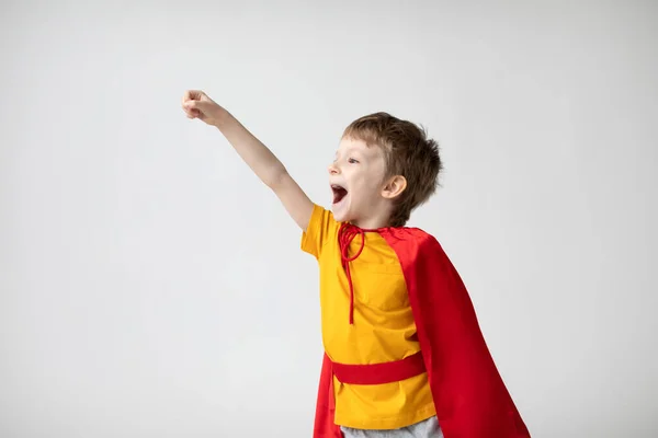 白い壁の背景に手を上げて赤いマントを着たスーパーヒーローの衣装を着た小さな男の子を笑っています 幸福と自由の概念 — ストック写真