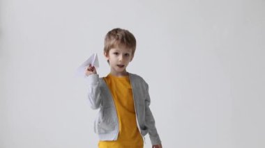 Sevimli küçük çocuk beyaz arka planda uçmak için kağıttan bir uçak fırlattı. Mutluluk ve olumlu duyguyu ifade ediyor. Yavaş çekim..