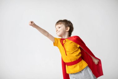 Kırmızı pelerinli gülümseyen küçük süper kahraman beyaz arka planda uçtuğunu gösteriyor. Güç kavramı. Küçük çocuk evde eğleniyor..