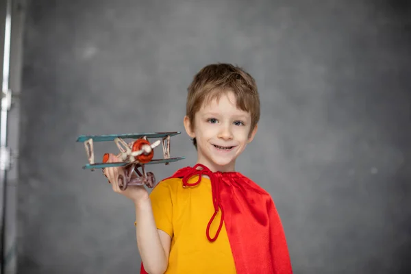カメラを見る肖像赤いケープの夢のような少年は 灰色の芸術的背景のおもちゃの飛行機で遊んでいます 小さなスーパーヒーロー — ストック写真