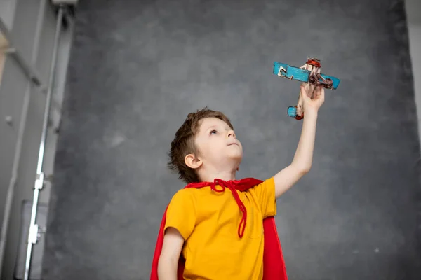 穿着红色斗篷的梦游男孩在灰色艺术背景下玩飞机玩具 小超级英雄 — 图库照片