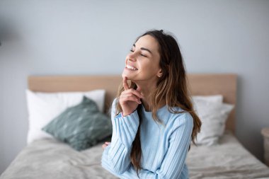 Yatakta oturmuş gülümseyen, sıradan giyinmiş hoş bir kadın. Rahat ev hayatı.
