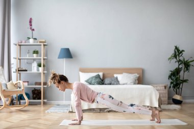Genç bir kadın tahta duruşunda duruyor ya da evde yoga minderinde şınav çekiyor. İçerde çalışıyor..