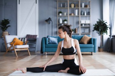 Oturma odasında yoga minderinde otururken evde egzersiz yapan genç ve sportif bir kadın. Ev çalışması.