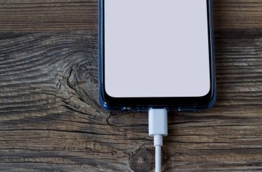 Ahşap yüzey üzerinde beyaz boş ekranlı akıllı telefon şarj kablosu takılı.