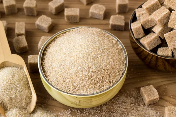 Doğal, kahverengi granüllü şeker bambu kasesinde kahverengi şeker küpleri ve tahta yüzeyde tahta kürek.