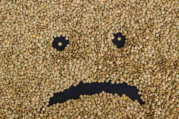 Sad face emoji with dry green lentils on black surface, harvest concept