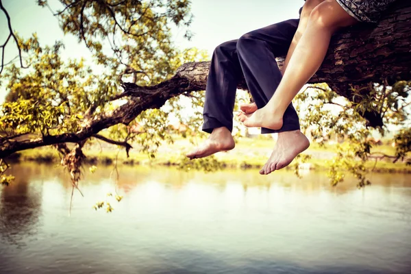 Young couple in love sitting cross-legged on a tree branch above river — Stock Photo, Image