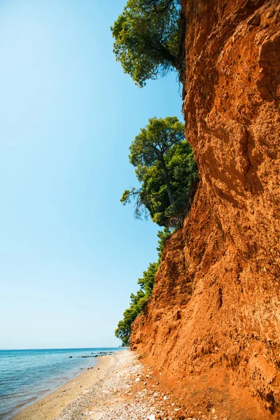 Coast of Aegean Sea with red ground and green pine trees — Stock Photo, Image