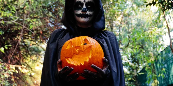 Young woman with halloween paint face mask wearing black hood - Scary witch holding spookey craved pumpkin