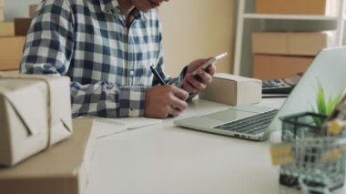 İnternet işletme sahibi telefonundaki mesajı kontrol ediyor ve detayları kağıda yazıyor ve teslimatı beklemek için ürüne yapışıyor. Online satış yönetim maliyetlerini düşürür. çevrimiçi gelenek kavramı