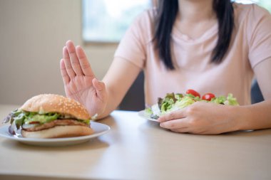 Sağlıklı Asyalı kadın bir hamburger tabağı itiyor ve salata yemeyi seçiyor. Yağ ve atıştırmalıkları diyet planına göre trans yağlarla ve temiz yiyeceklerle reddet..