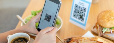 Kadının eli, restoranda bir QR kodunu taramak için telefon kullanıyor. İndirim almak ya da yemek için ödeme yapmak için. Para aktarmak için telefon kullan ya da internetten nakit ödeme yap. Pankart