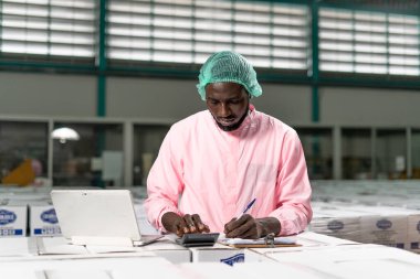 Amerikalı hammadde müfettişi fabrikada kullanılacak verileri not etmek için bilgisayarla çalışıyor. Depo çalışanları dizüstü bilgisayardan malzeme listesine bakıyor. Tedarikçiden hammadde al.