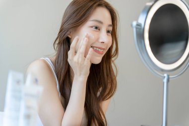 Güzel bir Asyalı kadın aynanın önünde oturuyor. Krem sürerek makyaj yapan sağlıklı bir kadın yüzü. Cilt kremi reklamı, kırışıklık önleyici krem, bebek yüzü.