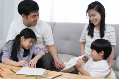 Asyalı bir aile evin içindeki kanepede gülüp eğleniyor. Baba ve anne sevgili oğluna ve kızına resim çizmeyi öğretiyor. Tatil ve aile mutluluğu.