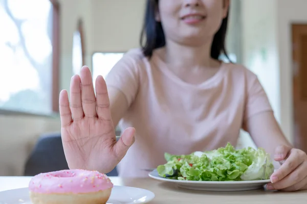Skinny asian woman pushes a donut plate and choose to eat salads. Refuse sweets and snacks with trans fats according to a diet plan and clean food.