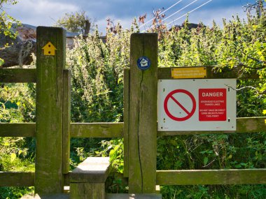Northumberland Sahil Yolu 'ndaki bir çit üzerindeki kırmızı ve beyaz uyarı işareti, yürüyenleri elektrik hatlarının tehlikesi konusunda uyarıyor. Yol yön işaretleri de görünür.