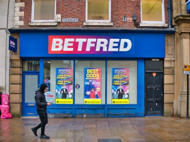 İngiltere 'nin kuzeyindeki Lancashire, Preston' daki Fishergate 'de bulunan ana cadde üzerindeki Betfred bahis zincirinin bir kolu. Genç bir insan yağmur yağarken yanından geçiyor.
