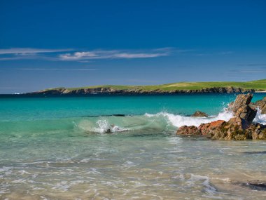 Dalgalar İngiltere 'nin güney Shetland kentindeki Scousburgh yakınlarında açık turkuaz bir sahilde kırılıyor. Güneşli bir günde, mavi bir gökyüzünde çekildi..