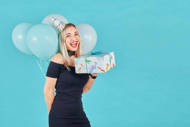 Siyah elbiseli ve doğum günü şapkalı güzel bir kadın elinde hediye kutusu ve mavi arka planda pastel hava balonları tutuyor. Mutlu genç kız doğum günü partisini kutluyor.