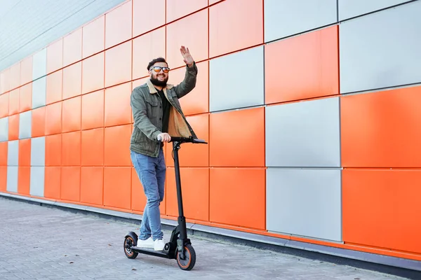Bearded man in sunglasses posing on electric scooter over orange wall background. Man riding kick scooter. — Stock Photo, Image