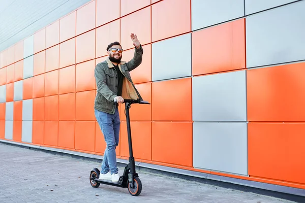 Brodatego mężczyznę w okularach przeciwsłonecznych pozującego na skuterze elektrycznym na pomarańczowym tle ściany. Człowiek jazda kick skuter. — Zdjęcie stockowe