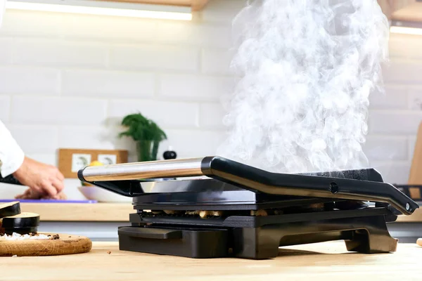 The woman\'s hands prepares meat on an electric grill on wooden table. Process of cooking meat on an electric grill. Smoke in the home kitchen.