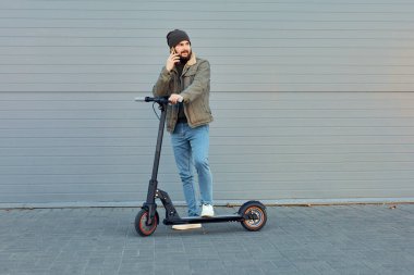 Duvarın yanında elektrikli scooterlı adam. Ekolojik ulaşım konsepti. Şık sakallı erkek telefon kullanıyor, e-scooter 'da poz veriyor..