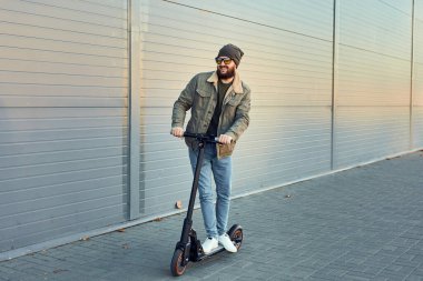 Duvarın yanında elektrikli scooterlı adam. Ekolojik ulaşım konsepti. Güneş gözlüklü şık erkek e-scooter 'da poz veriyor..
