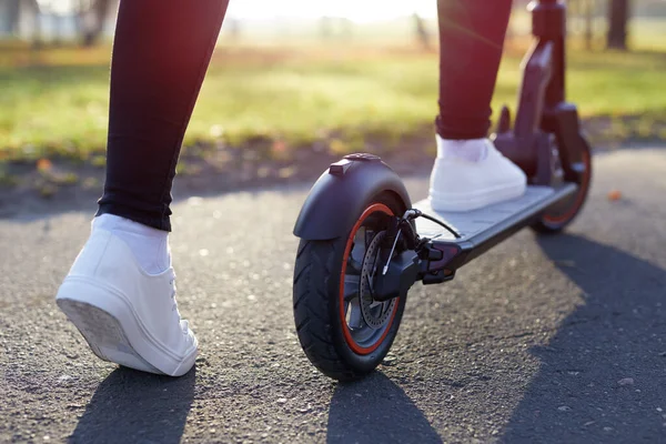 若い女性は 電動スクーターや電子スクーターで日没で街や公園を発見します スポーツスニーカーの女性の足は電動スクーターに立つ 生態系と都市交通に乗って女の子 — ストック写真