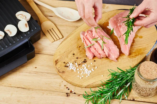 キッチンで肉ステーキを作る女性 女性シェフはローズマリーと塩でキッチンの背景に新鮮な生肉を振りかけます 木製のテーブルの上の電気グリルと肉 ストック画像