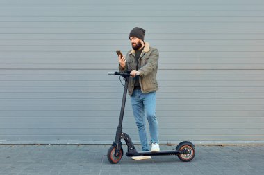 Duvarın yanında elektrikli scooterlı adam. Ekolojik ulaşım konsepti. Şık sakallı erkek telefon kullanıyor, e-scooter 'da poz veriyor..